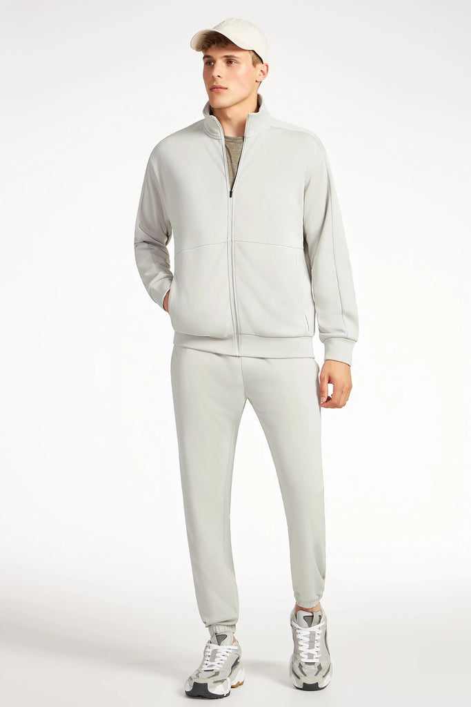 A man models the TECH APPAREL MENS NEO-KNIT ZIP-UP MOCK NECK SWEATER in light gray with matching pants, beige cap, and gray sneakers. One hand in his pocket, he looks aside against a plain white background.