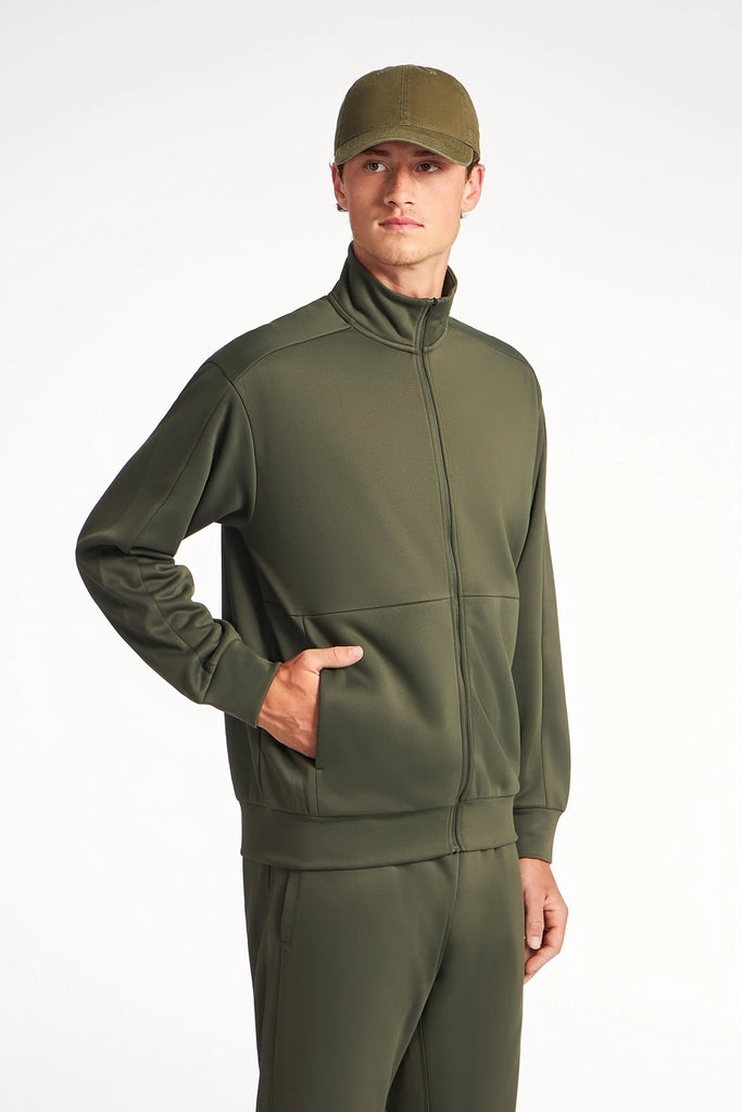 A man in a TECH APPAREL MENS NEO-KNIT ZIP-UP MOCK NECK SWEATER and matching cap stands against a white background, one hand in his pocket, gazing to the side.