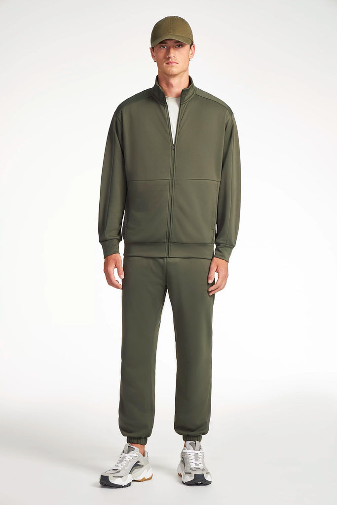 A man faces forward, wearing a TECH APPAREL MENS NEO-KNIT ZIP-UP MOCK NECK SWEATER and matching joggers in olive green, paired with a white T-shirt, white sneakers, and an olive green cap against a plain white background.