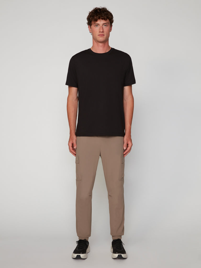 A young man with curly hair stands facing forward against a plain background, wearing a black t-shirt, black sneakers, and TECH APPAREL MEN’S FLUX 2.0 RELAXED FIT PULL-ON CARGO JOGGERS in beige. His arms are relaxed at his sides.