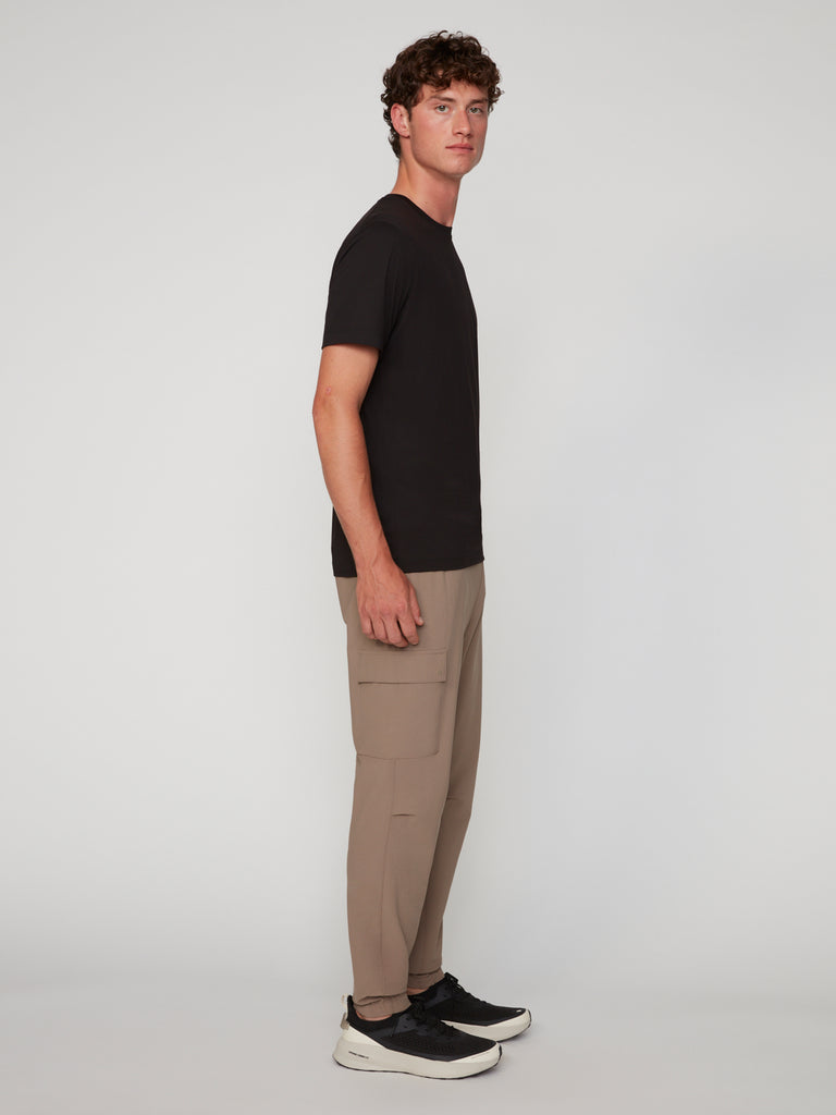 A young man stands sideways in a plain light gray studio, wearing a black t-shirt, TECH APPAREL MEN’S FLUX 2.0 RELAXED FIT PULL-ON CARGO JOGGER in beige, and black sneakers with white soles.