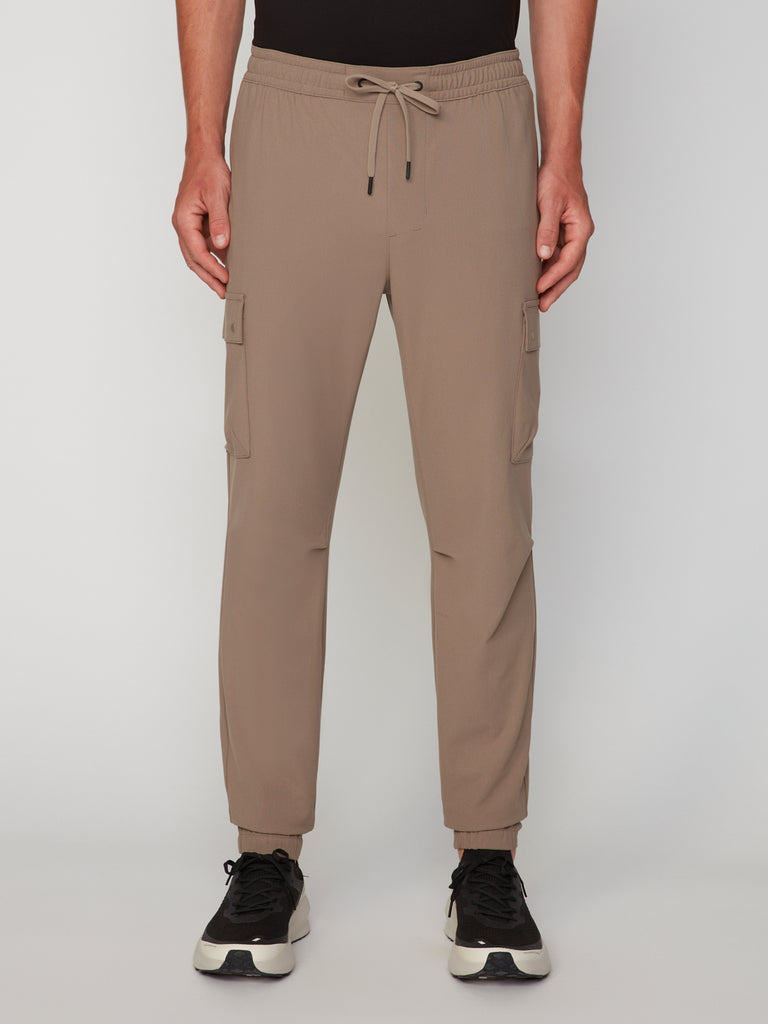 A man wears TECH APPAREL MEN’S FLUX 2.0 RELAXED FIT PULL-ON CARGO JOGGER in taupe stretch fabric with side pockets and a drawstring waist, styled with black and white sneakers, standing against a plain white background.