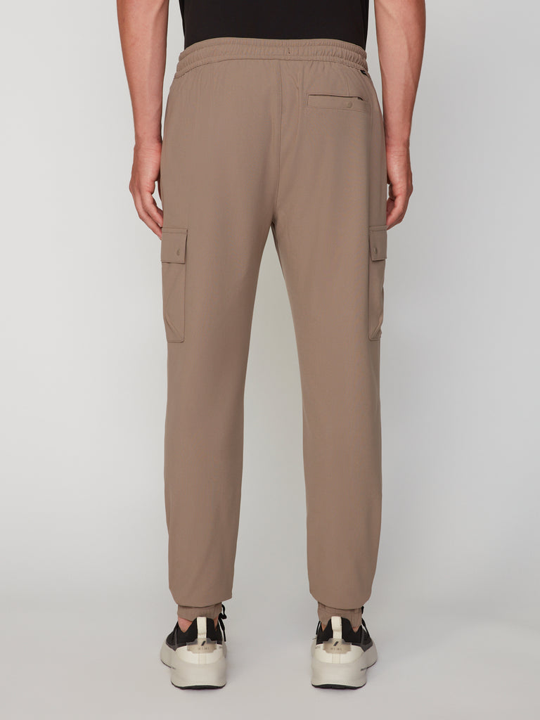 A person is shown from the back wearing TECH APPAREL’s MEN’S FLUX 2.0 RELAXED FIT PULL-ON CARGO JOGGER in light brown with side pockets and a right zipper pocket, paired with white sneakers, against a plain light background.