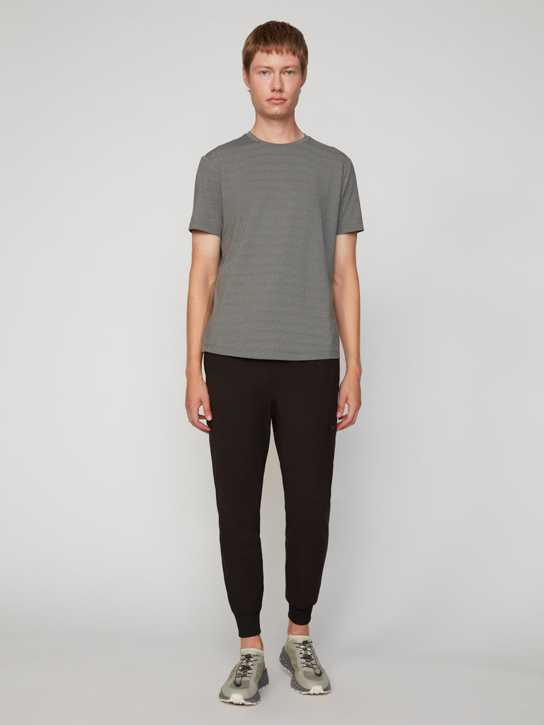 A person stands facing forward against a plain light background, wearing a gray short-sleeve t-shirt, gray sneakers, and TECH APPAREL MEN’S FLUX 2.0 RELAXED FIT PULL-ON CARGO JOGGER in black with stretch fabric. Their arms are relaxed by their sides.