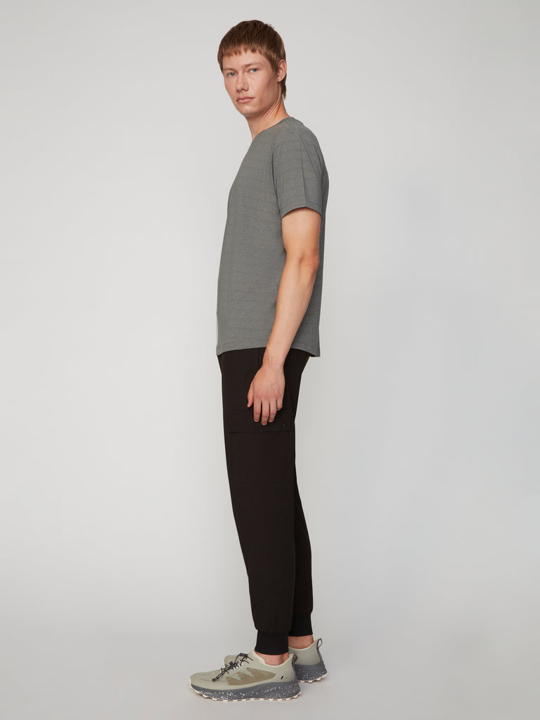 A person with short light brown hair stands sideways against a light gray background, wearing a gray t-shirt, light gray sneakers, and the TECH APPAREL MEN’S FLUX 2.0 RELAXED FIT PULL-ON CARGO JOGGER in black stretch fabric.