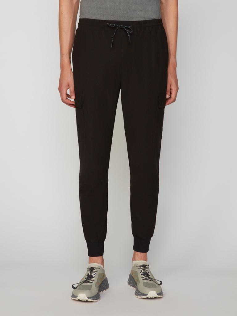 A person wears TECH APPAREL’s MEN’S FLUX 2.0 RELAXED FIT PULL-ON CARGO JOGGER featuring stretch fabric, drawstring waist, side cargo pockets, and cuffed ankles with a gray fitted shirt and beige sneakers against a plain light background.