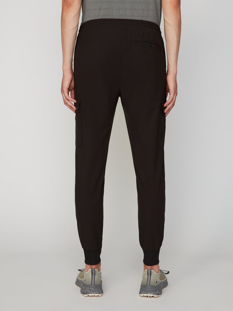 A person stands facing away, wearing TECH APPAREL MEN’S FLUX 2.0 RELAXED FIT PULL-ON CARGO JOGGER pants with side and back pockets, a gray shirt, and gray athletic shoes against a plain light background.