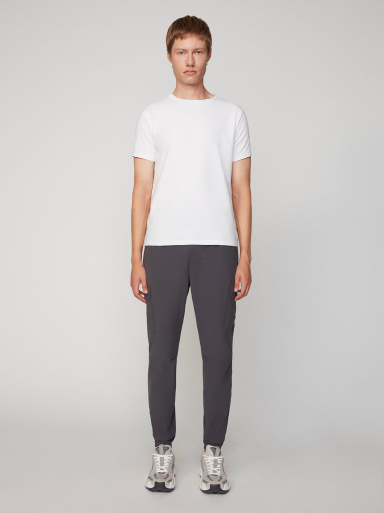 A person stands facing forward, wearing a plain white short-sleeve t-shirt, TECH APPAREL MEN’S FLUX 2.0 RELAXED FIT PULL-ON CARGO JOGGER in dark grey stretch fabric, and grey sneakers against a plain light grey background.