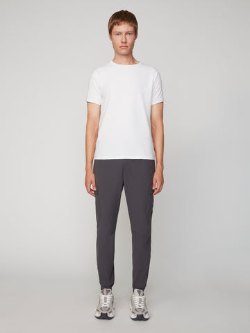 A person stands facing forward, wearing a plain white short-sleeve t-shirt, TECH APPAREL MEN’S FLUX 2.0 RELAXED FIT PULL-ON CARGO JOGGER in dark grey stretch fabric, and grey sneakers against a plain light grey background.