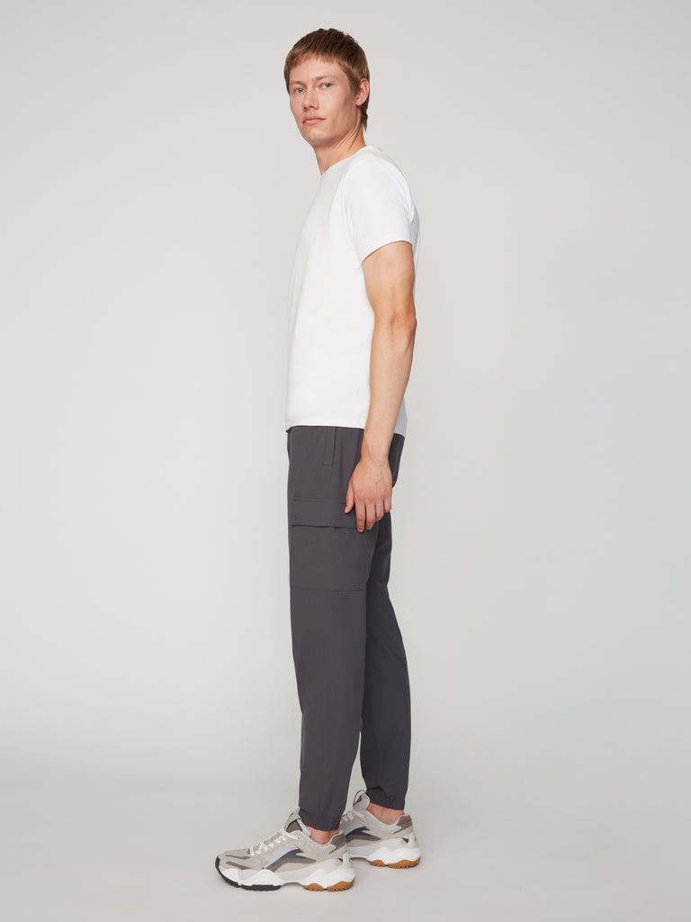 A man stands sideways wearing a white t-shirt, gray sneakers, and TECH APPAREL MEN’S FLUX 2.0 RELAXED FIT PULL-ON CARGO JOGGER pants made from stretch fabric against a plain light background.