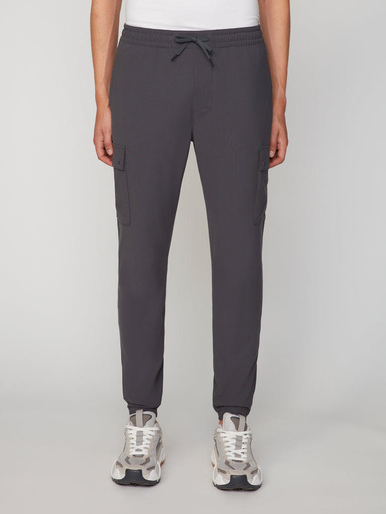 A model wears TECH APPARELs MEN’S FLUX 2.0 RELAXED FIT PULL-ON CARGO JOGGER in dark gray with white sneakers and a white top, standing against a light gray background, highlighting modern men’s activewear style.