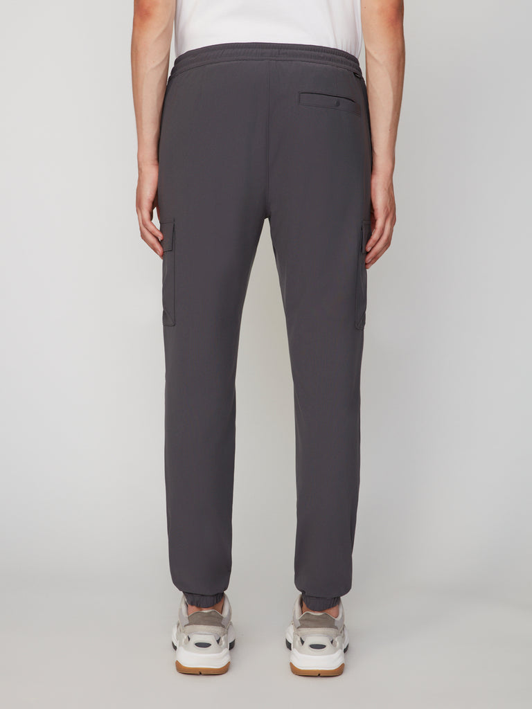 A person faces away from the camera, wearing TECH APPAREL MEN’S FLUX 2.0 RELAXED FIT PULL-ON CARGO JOGGER in dark gray with side pockets, paired with a white T-shirt and white sneakers, set against a plain light background.