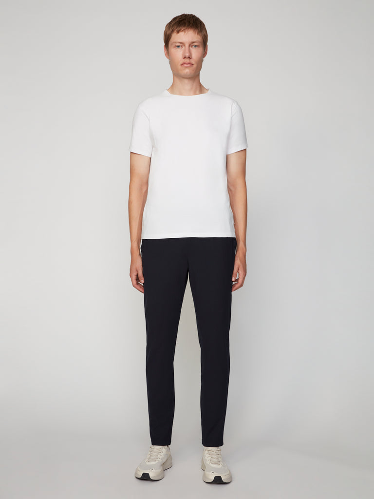 A person stands facing forward against a plain light background, wearing a white short-sleeve t-shirt, TECH APPAREL MEN’S FLUX 2.0 RELAXED FIT PULL-ON CHINO PANT in black, and white sneakers.