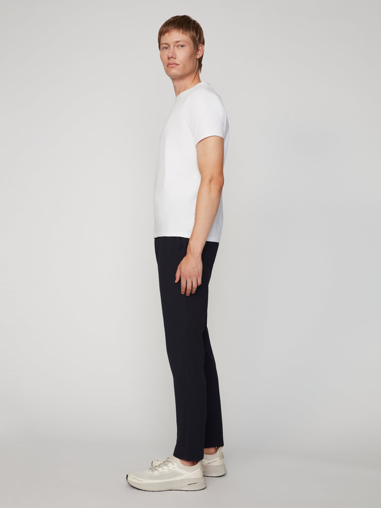 A man stands sideways facing left, wearing a white t-shirt, TECH APPAREL MEN’S FLUX 2.0 RELAXED FIT PULL-ON CHINO PANT, and white sneakers against a plain light gray background.