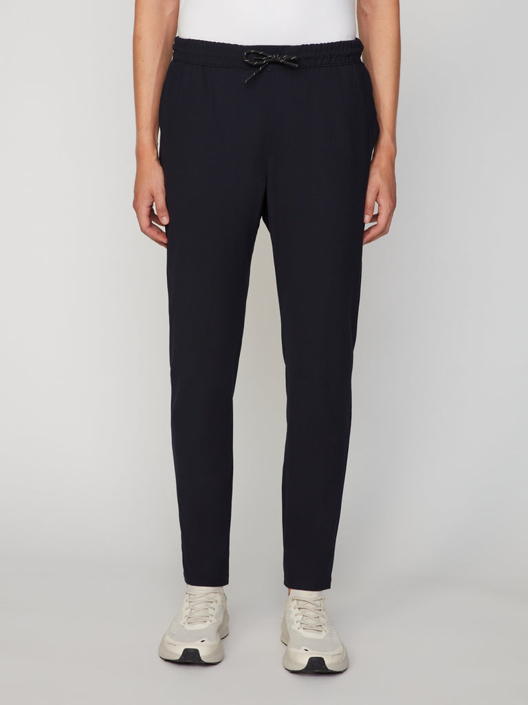 A person wears TECH APPAREL MEN’S FLUX 2.0 RELAXED FIT PULL-ON CHINO PANT in black with white sneakers, standing against a plain light gray background, hands relaxed by their sides. The upper body is partially out of frame.