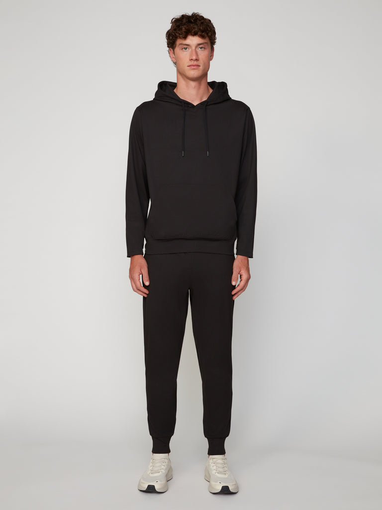 A person faces forward wearing TECH APPAREL’s MEN’S TECH:SOFT CLASSIC FIT PULL-ON CHINO JOGGER in black with a matching hoodie and white sneakers, set against a plain, light gray background.