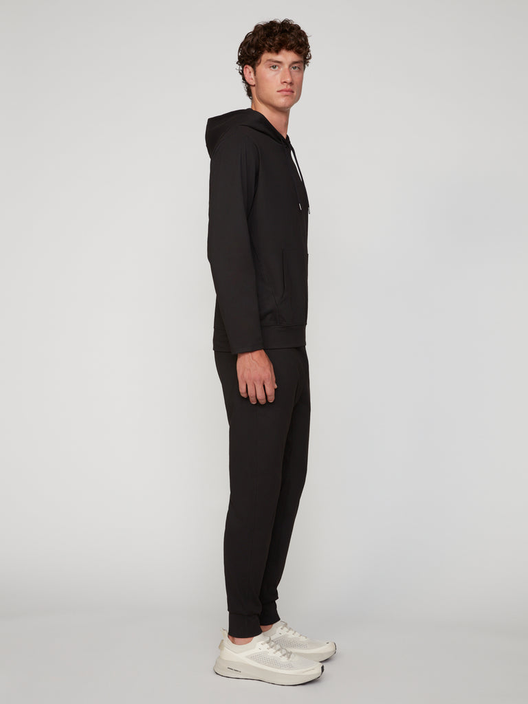 A person in profile faces left, wearing a black hooded sweatshirt, TECH APPAREL MEN’S TECH:SOFT CLASSIC FIT PULL-ON CHINO JOGGER pants, and white sneakers against a plain light background.