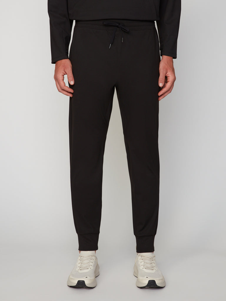 A person wearing TECH APPAREL MEN’S TECH:SOFT CLASSIC FIT PULL-ON CHINO JOGGER pants in black, paired with a matching long-sleeve top and white sneakers, stands against a plain light gray background. Upper body is mostly out of frame.