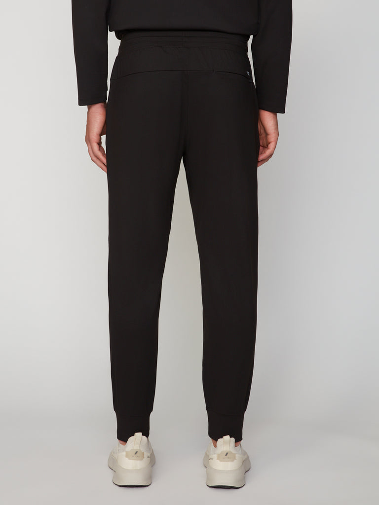 A person is shown from the back wearing TECH APPAREL MEN’S TECH:SOFT CLASSIC FIT PULL-ON CHINO JOGGER pants in black with cuffed ankles and white sneakers, standing against a plain light gray background. The joggers feature relaxed fit and Tech:Soft fabric.