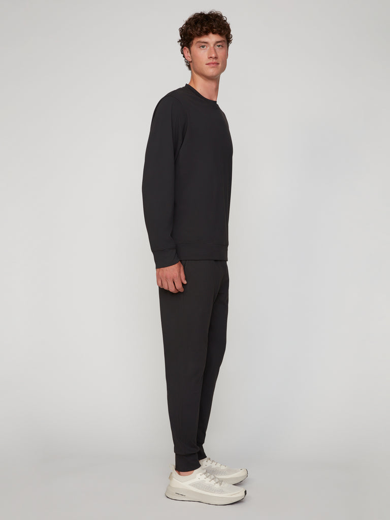 A man with short curly hair stands sideways against a light gray background, wearing a black long-sleeve sweatshirt, white sneakers, and TECH APPAREL MEN’S TECH:SOFT CLASSIC FIT PULL-ON CHINO JOGGER pants.