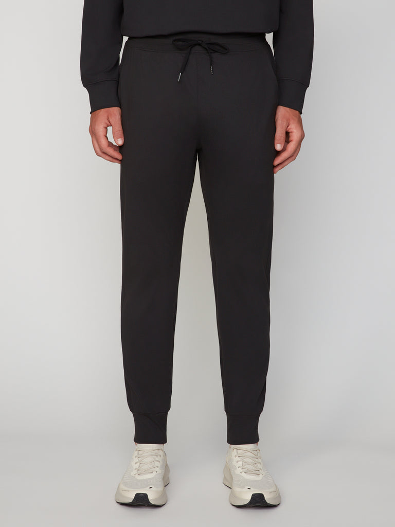 A person wears TECH APPAREL MEN’S TECH:SOFT CLASSIC FIT PULL-ON CHINO JOGGER pants with a drawstring waist and white sneakers, standing against a plain light background.