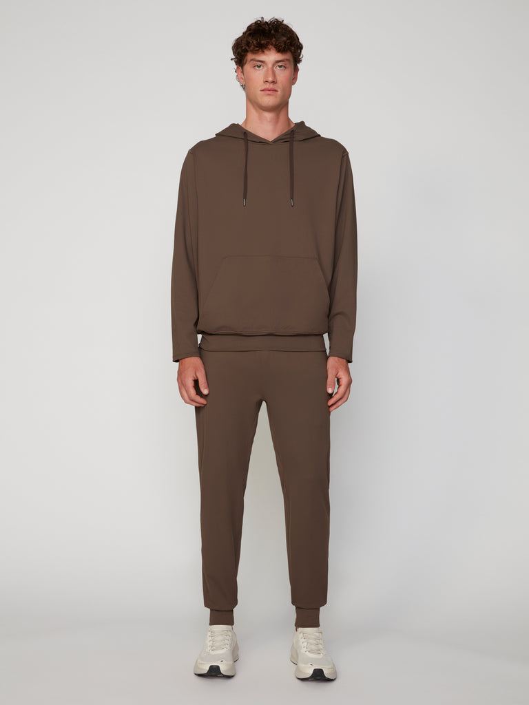 A young man faces forward in a brown TECH APPAREL MEN’S TECH:SOFT CLASSIC FIT PULL-ON CHINO JOGGER and matching hooded sweatshirt, paired with white sneakers against a plain, light background.
