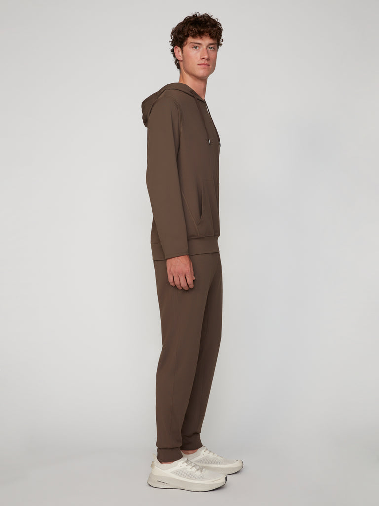 A young man stands sideways in TECH APPAREL’s MEN’S TECH:SOFT CLASSIC FIT PULL-ON CHINO JOGGER, paired with white sneakers, against a plain light gray background.