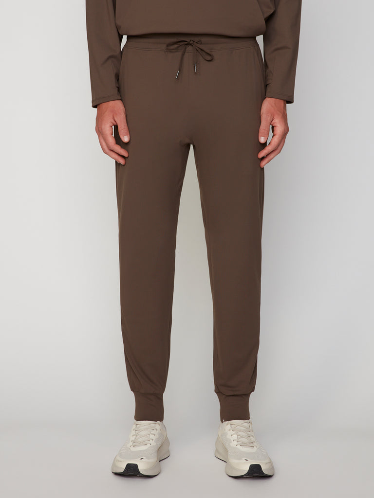 A person wearing TECH APPAREL MEN’S TECH:SOFT CLASSIC FIT PULL-ON CHINO JOGGER and white sneakers stands against a plain light background; the photo is cropped from shoulders to feet.