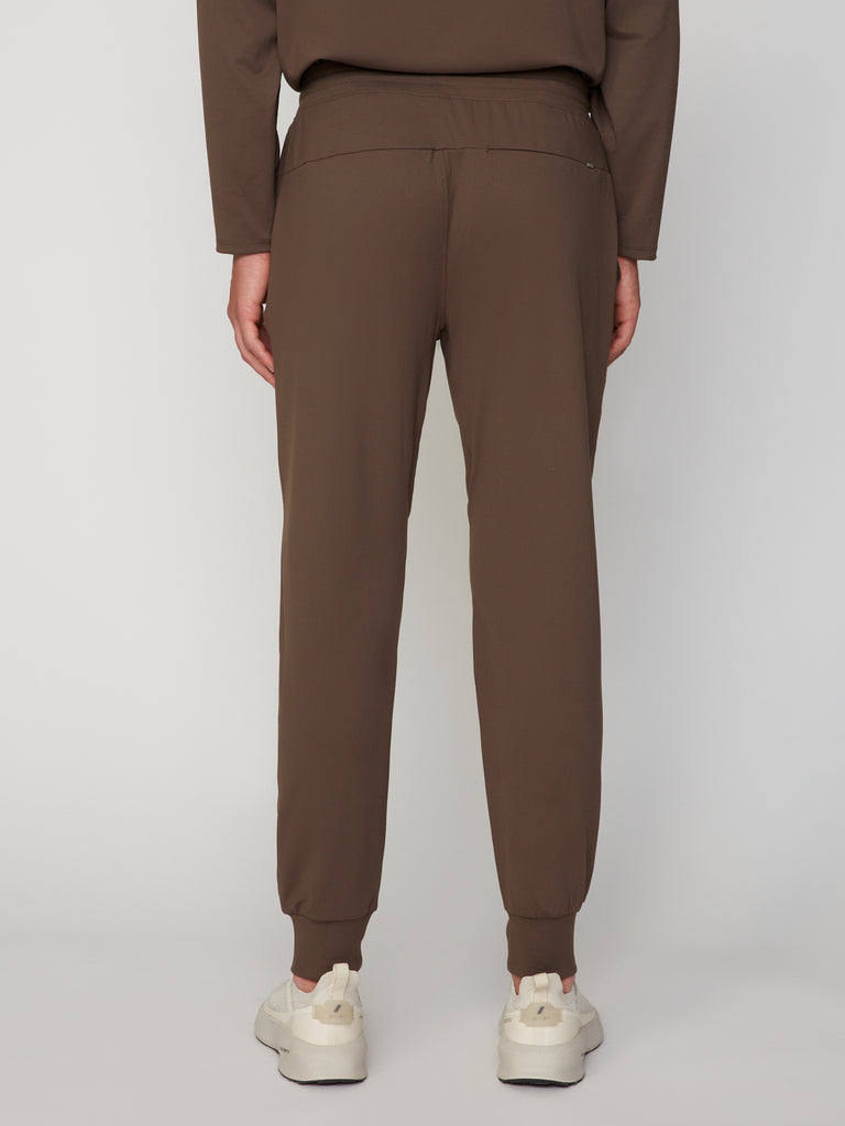 A person faces away from the camera, wearing TECH APPAREL’s MEN’S TECH:SOFT CLASSIC FIT PULL-ON CHINO JOGGER in brown, paired with a matching top and white sneakers against a plain light background.