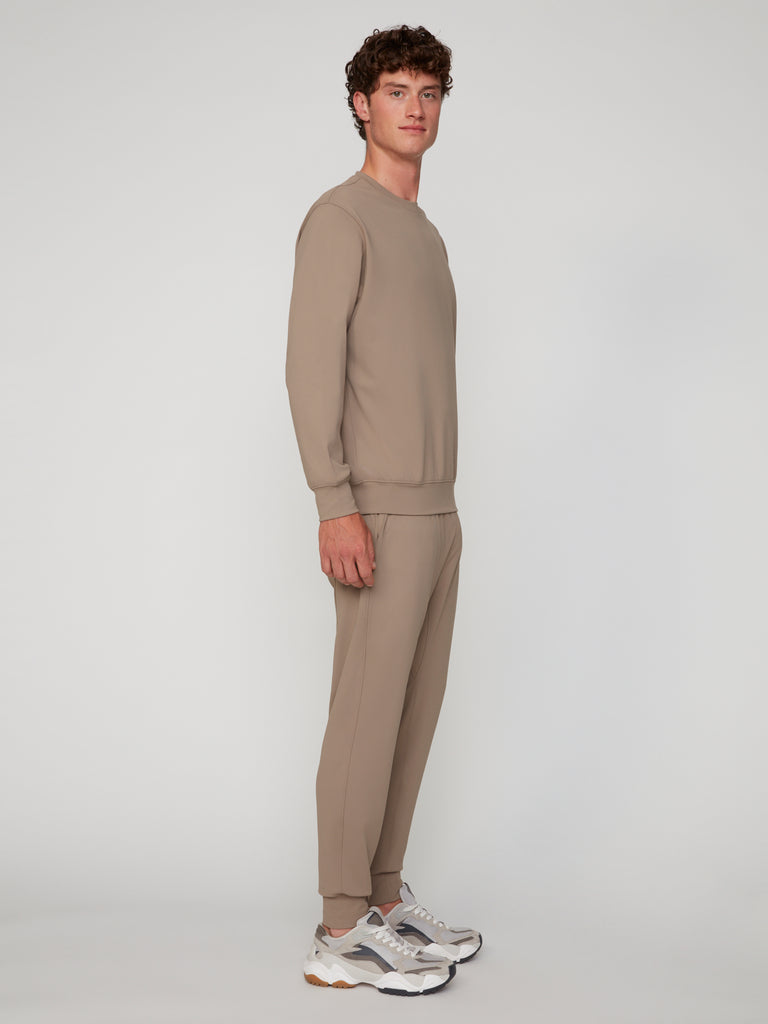 A young man stands sideways in TECH APPAREL’s MEN’S TECH:SOFT CLASSIC FIT PULL-ON CHINO JOGGER and a matching sweatshirt, paired with white and gray sneakers, against a plain light background.