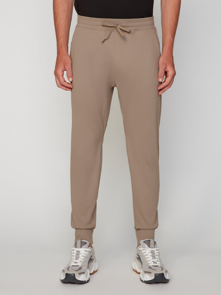 A person wears TECH APPAREL MEN’S TECH:SOFT CLASSIC FIT PULL-ON CHINO JOGGER pants—tan, with a drawstring waist and cuffed ankles—paired with gray-and-white sneakers, standing against a plain white background.