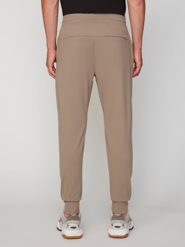 A person is seen from the back against a plain light background, wearing TECH APPAREL MEN’S TECH:SOFT CLASSIC FIT PULL-ON CHINO JOGGER pants with a zip pocket and white sneakers. These casual pants offer comfort thanks to tech:soft fabric.