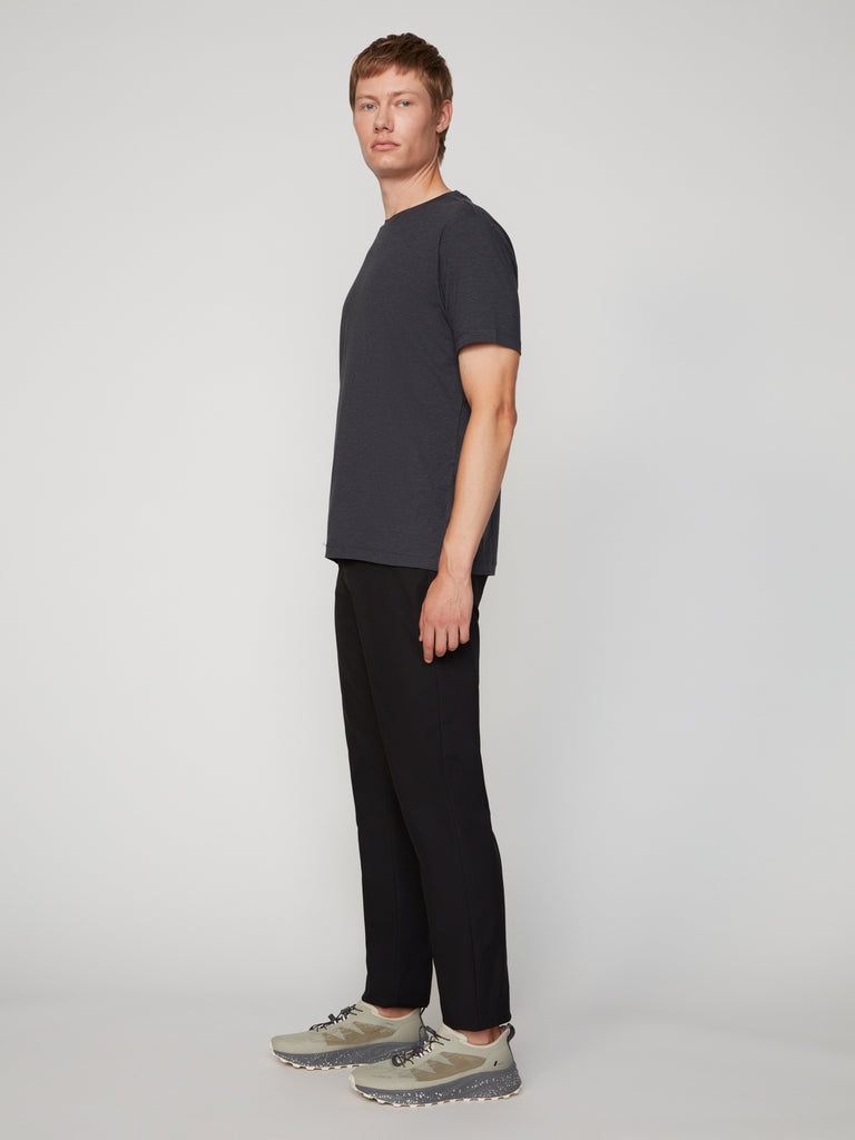 A man with short light brown hair stands sideways in a dark gray t-shirt and TECH APPAREL MEN’S 247:TWILL RELAXED TAPERED FIT 5-POCKET PANT, paired with light gray sneakers, against a plain light background.