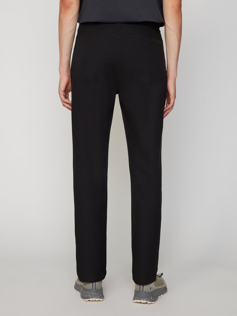 A person faces away from the camera against a light gray background, wearing TECH APPAREL MEN’S 247:TWILL RELAXED TAPERED FIT 5-POCKET PANT, a dark shirt, and gray lace-up sneakers.