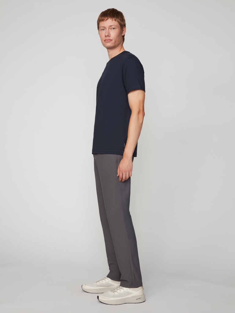 A man stands relaxed, facing sideways, wearing a navy blue t-shirt, TECH APPAREL MEN’S 247:TWILL RELAXED TAPERED FIT 5-POCKET PANT in gray, and white sneakers against a plain, light-colored background.