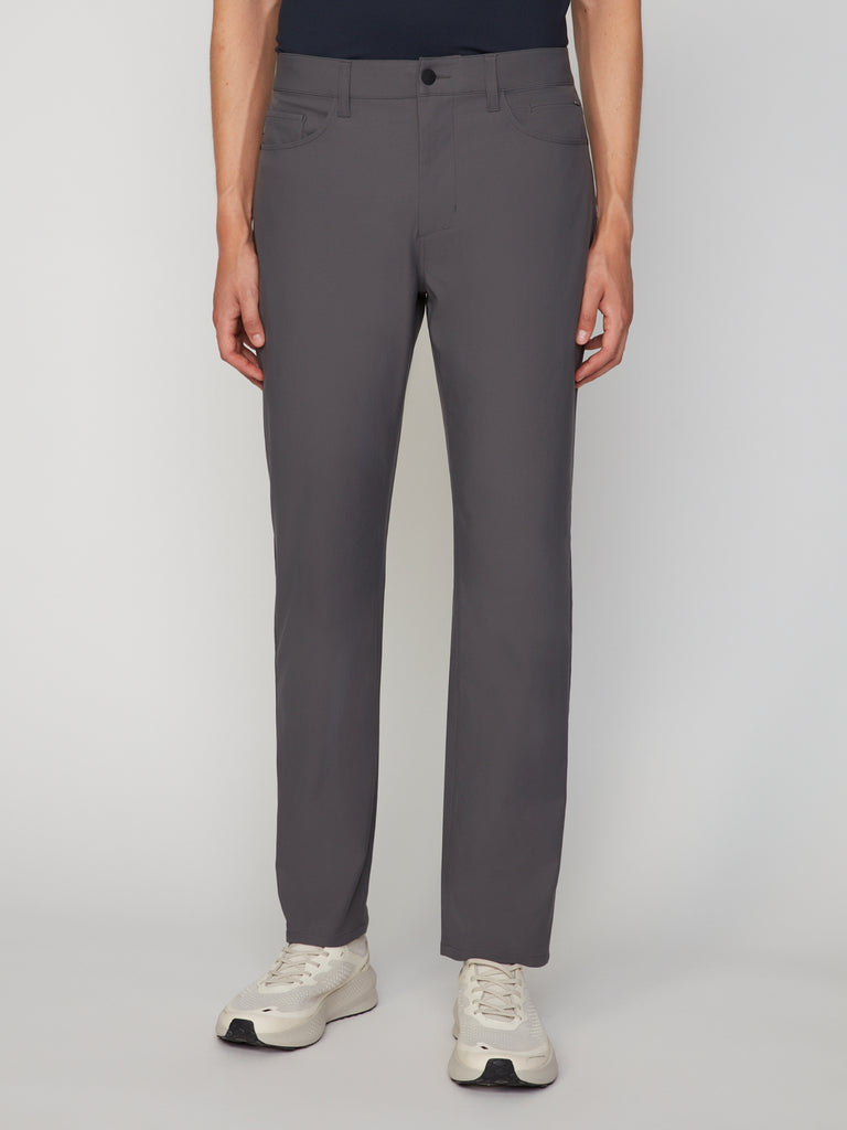 A person in TECH APPAREL MEN’S 247:TWILL RELAXED TAPERED FIT 5-POCKET PANT, dark top, and white sneakers stands against a light background, shown from shoulders to feet with hands at their sides.