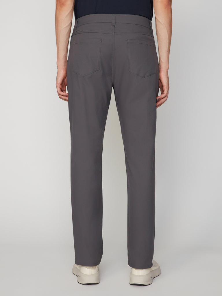 A man wearing TECH APPAREL MEN’S 247:TWILL RELAXED TAPERED FIT 5-POCKET PANT in gray with light-colored shoes, seen from the back against a plain white background.