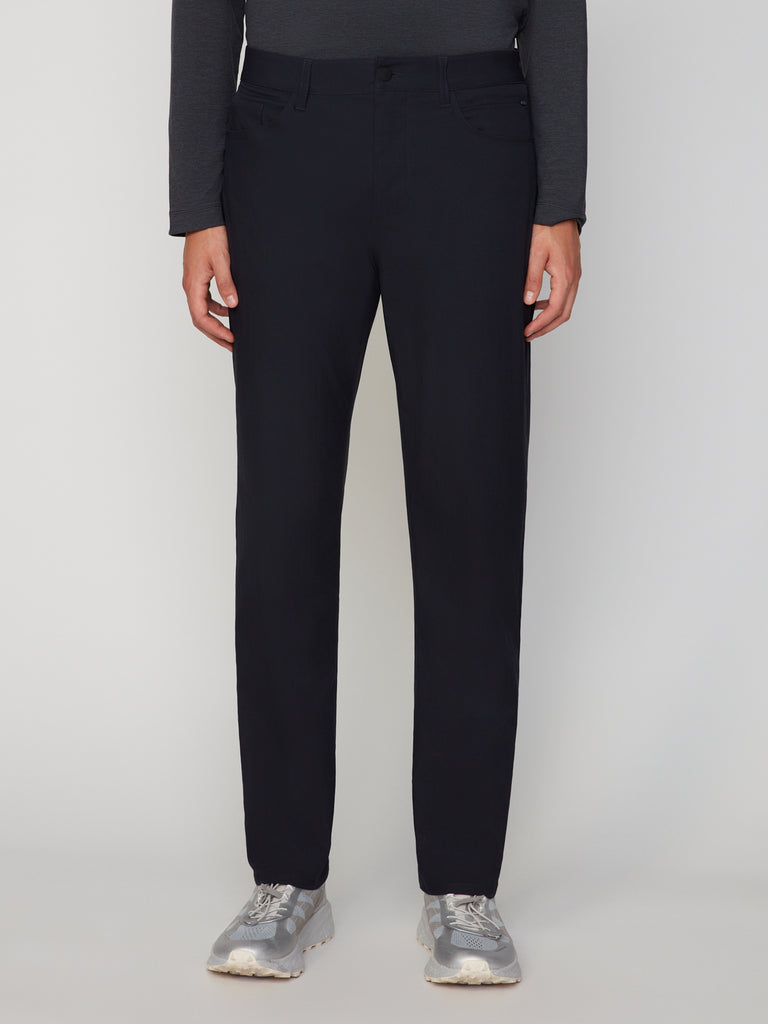 Wearing TECH APPAREL’s MEN’S 247:TWILL RELAXED TAPERED FIT 5-POCKET PANT in black, paired with a dark long-sleeve shirt and silver sneakers, a person stands against a plain light background, showing only their lower torso, legs, and feet.