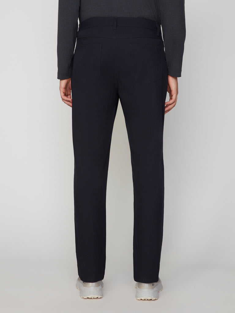 A person faces away from the camera, wearing TECH APPAREL MEN’S 247:TWILL RELAXED TAPERED FIT 5-POCKET PANT in black, a dark long-sleeve shirt, and light gray sneakers against a plain light background.