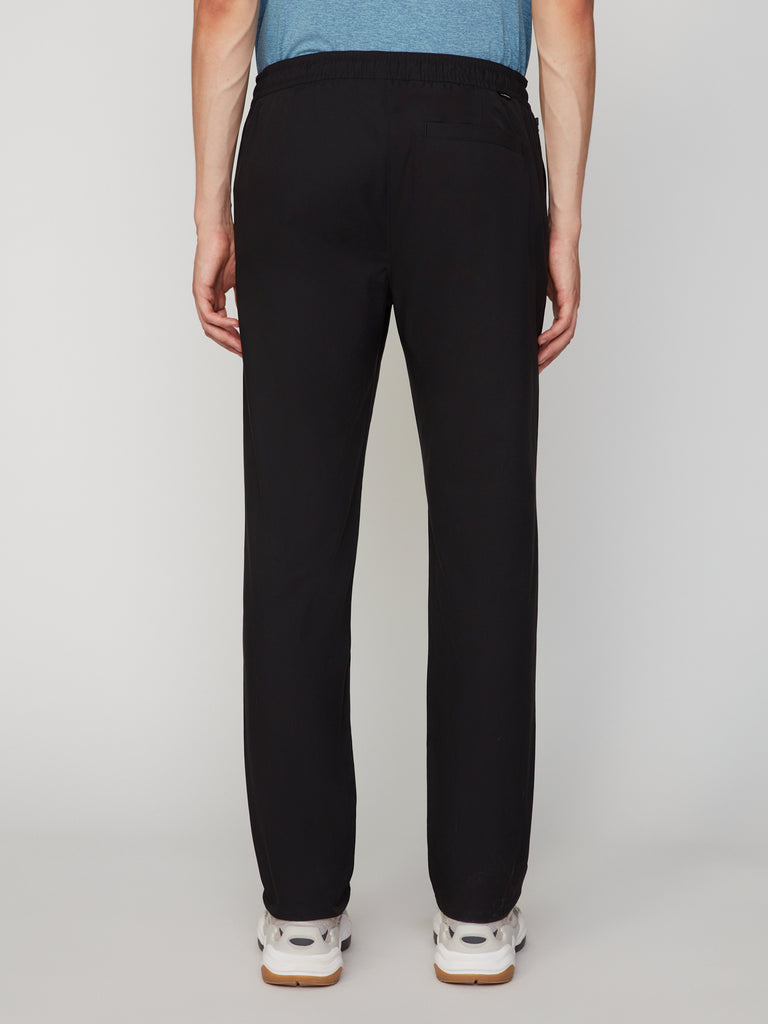 A man is shown from the back in TECH APPAREL MENS PULL-ON CHINO 247:TWILL PANT, relaxed straight fit black pants with an elastic waistband and single back pocket, paired with white sneakers, against a plain light background.