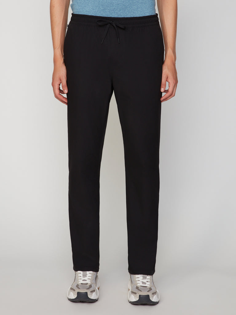 A person wears TECH APPAREL MENS PULL-ON CHINO 247:TWILL PANT in black with a relaxed straight fit, paired with a blue shirt and white athletic shoes. Only the lower half is shown, standing against a plain light background.