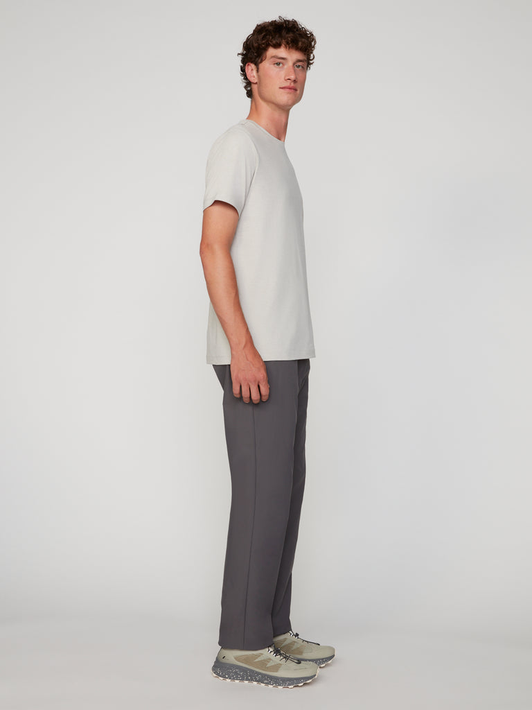 A young man stands in profile against a plain light background, wearing a light grey short-sleeve t-shirt, TECH APPAREL MENS PULL-ON CHINO 247:TWILL PANT in dark grey, and beige sneakers with a textured sole.