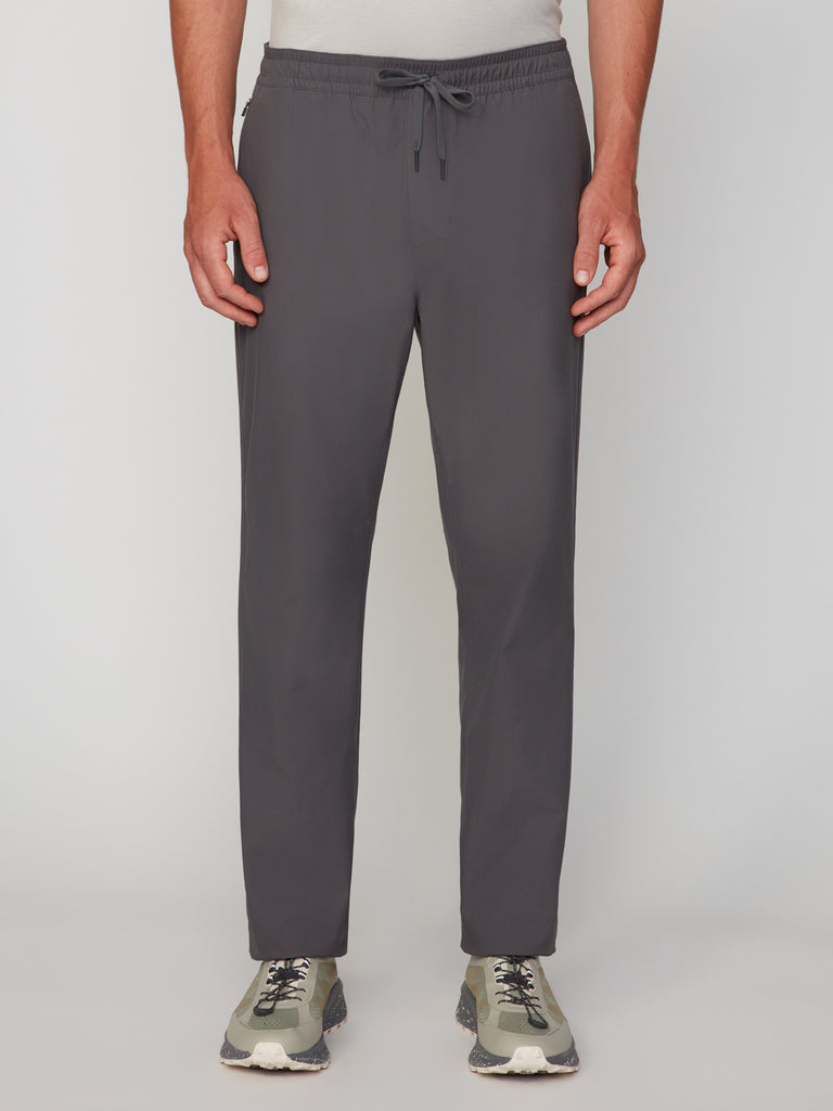 A person in TECH APPARELs MENS PULL-ON CHINO 247:TWILL PANT, dark gray with a relaxed straight fit and mechanical stretch twill, pairs it with a light shirt and light green sneakers, standing against a plain white background.