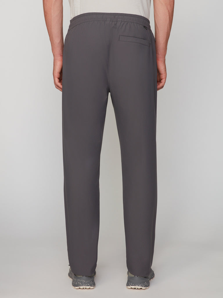 A person wearing TECH APPAREL MENS PULL-ON CHINO 247:TWILL PANT in dark gray mechanical stretch twill and gray shoes is shown from the back. The pants feature an elastic waistband and a right-side back pocket.