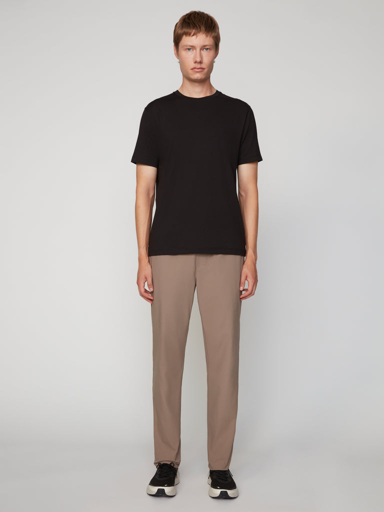 A person stands facing forward against a light-gray background, wearing a black short-sleeve t-shirt, TECH APPAREL MENS PULL-ON CHINO 247:TWILL PANT in taupe with relaxed straight fit, and black sneakers with white soles.