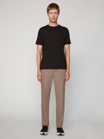 A person stands facing forward against a light-gray background, wearing a black short-sleeve t-shirt, TECH APPAREL MENS PULL-ON CHINO 247:TWILL PANT in taupe with relaxed straight fit, and black sneakers with white soles.