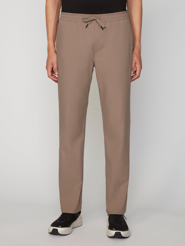 A person wears TECH APPARELs MENS PULL-ON CHINO 247:TWILL PANT in taupe, relaxed straight fit, paired with black-and-white sneakers. The stretch twill pants are shown from shoulders down against a plain white background.