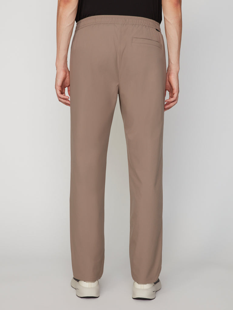 A person is seen from behind wearing TECH APPAREL MENS PULL-ON CHINO 247:TWILL PANT—relaxed straight fit with stretch twill, elastic waistband, single back pocket—paired with a black top and white shoes.