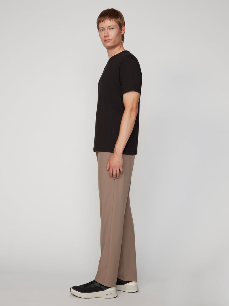 A person stands sideways in a black t-shirt and TECH APPAREL MENS PULL-ON CHINO 247:TWILL PANT in beige stretch twill, paired with black and white sneakers, looking toward the camera against a plain light gray background.