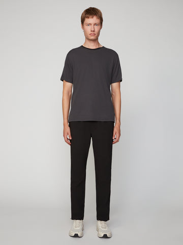 A young man stands against a light gray background, wearing TECH APPAREL’s MEN’S PULL-ON TRAINING PANT, RELAXED TAPERED FIT in performance stretch fabric, paired with a dark gray t-shirt and white sneakers. He faces forward with arms relaxed.